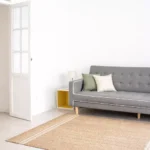A small sunlit living room featuring a natural jute rug and sage green pillows, showcasing summer 2026 rugs for small spaces.